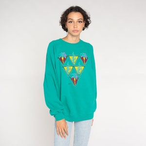 May include: Teal sweatshirt with a colorful geometric design. The design features a triangle of smaller triangles with palm tree and cactus illustrations. The sweatshirt has long sleeves and a crew neck. The model is wearing light blue jeans.