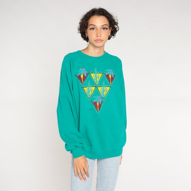 May include: Teal sweatshirt with a colorful geometric design. The design features a triangle of smaller triangles with palm tree and cactus illustrations. The sweatshirt has long sleeves and a crew neck. The model is wearing light blue jeans.