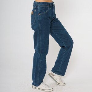 May include: Dark blue denim jeans with a classic five-pocket design. The jeans have a straight leg fit and a brown leather patch on the back pocket. The model is wearing white sneakers.