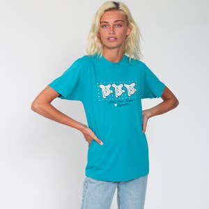 May include: A turquoise blue t-shirt with a graphic of three white bears wearing hats and the text "Head for Hills" with a heart and the word "Sygnature".