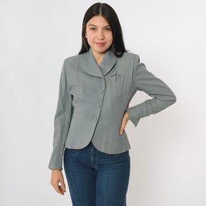 May include: A light gray blazer with a notched collar and three buttons. The blazer has a fitted silhouette and long sleeves. A small decorative detail is on the left side of the chest. The person is wearing blue jeans.