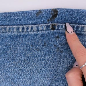 Pode incluir: Close-up de tecido jeans azul com manchas escuras perto da cintura. O detalhe da costura &eacute; vis&iacute;vel ao longo da costura. Um dedo com uma unha pintada aponta para a mancha. V&aacute;rios an&eacute;is est&atilde;o no dedo.