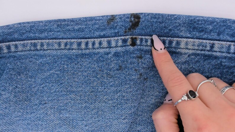 Pode incluir: Close-up de tecido jeans azul com manchas escuras perto da cintura. O detalhe da costura &eacute; vis&iacute;vel ao longo da costura. Um dedo com uma unha pintada aponta para a mancha. V&aacute;rios an&eacute;is est&atilde;o no dedo.