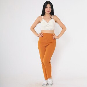 May include: A woman is wearing a white crochet top and mustard yellow high-waisted pants. She is standing on a white background.