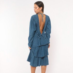 Puede incluir: Un vestido de mezclilla de manga larga con espalda descubierta y tiras finas para atar al cuello. El vestido incluye un lazo prominente en la cintura y una falda con volantes de varias capas que cae por debajo de las rodillas en azul medio.