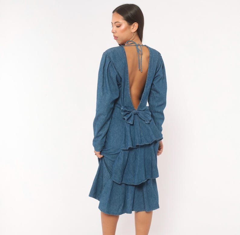 Puede incluir: Un vestido de mezclilla de manga larga con espalda descubierta y tiras finas para atar al cuello. El vestido incluye un lazo prominente en la cintura y una falda con volantes de varias capas que cae por debajo de las rodillas en azul medio.