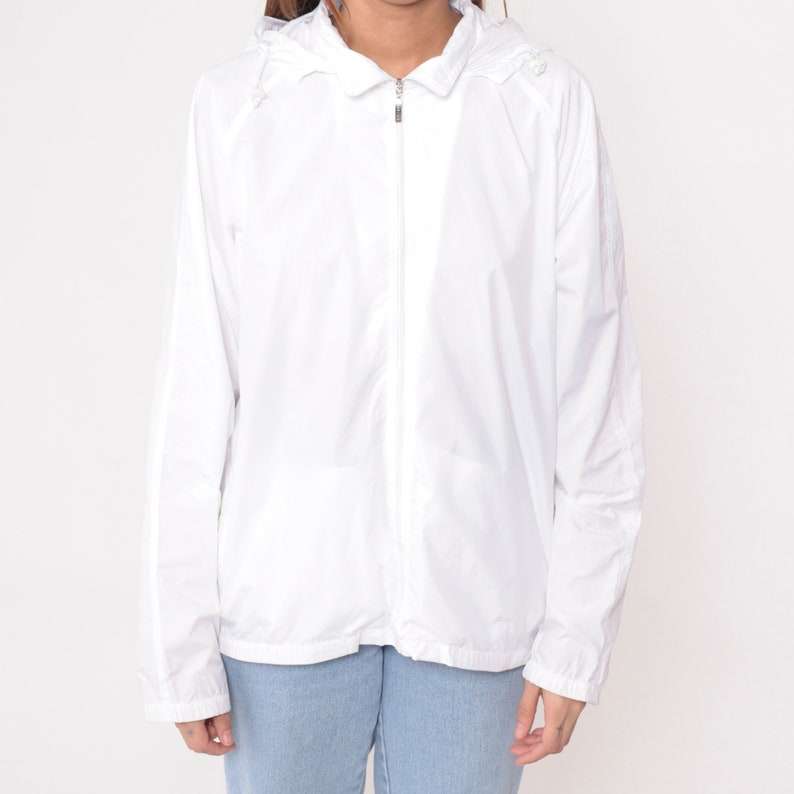 May include: A white hooded jacket with a full-length zipper closure. The jacket has long sleeves and a drawstring hood. The material appears lightweight and suitable for layering. The jacket is paired with light blue jeans.