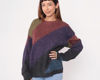 May include: A multi-colored sweater with a diagonal color block design. The sweater features shades of olive green, burgundy, purple, gray, and blue. The sweater is made of a textured knit material and has a crew neck.