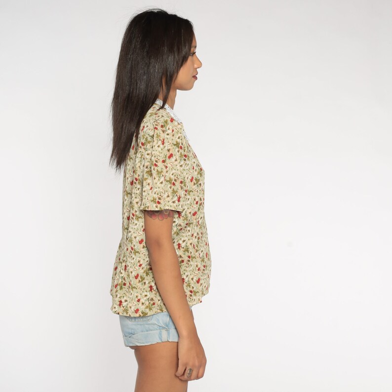 K&ouml;nnte beinhalten: Ein beiges, kurz&auml;rmeliges Oberteil mit Blumenmuster und roten und gr&uuml;nen Akzenten. Es hat einen wei&szlig;en Spitzenkragen und wird mit hellblauen Jeansshorts kombiniert. Das Blumenmuster wiederholt sich auf dem Stoff.