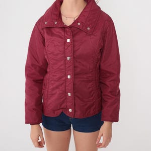 May include: A burgundy-colored jacket with a high collar and silver square buttons. The jacket has a quilted texture and a fitted waist. The sleeves are long, and the jacket appears to be made of a lightweight material. The jacket is paired with denim shorts.