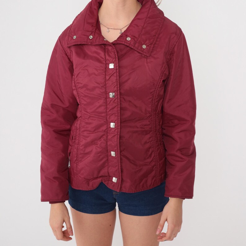 May include: A burgundy-colored jacket with a high collar and silver square buttons. The jacket has a quilted texture and a fitted waist. The sleeves are long, and the jacket appears to be made of a lightweight material. The jacket is paired with denim shorts.