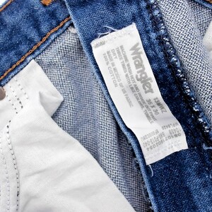 May include: Close-up of blue denim jeans with a white interior and a Wrangler label. The label indicates the jeans are size 35 x 36. The jeans have a classic five-pocket design and are made of cotton.