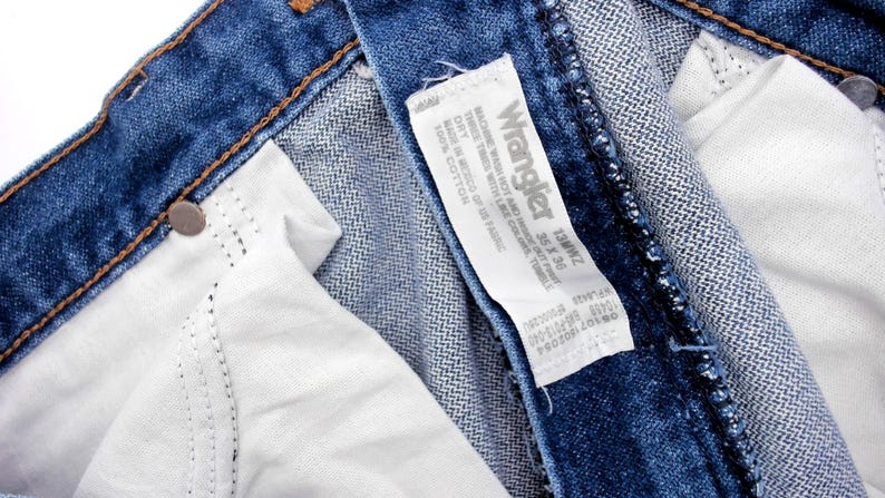 May include: Close-up of blue denim jeans with a white interior and a Wrangler label. The label indicates the jeans are size 35 x 36. The jeans have a classic five-pocket design and are made of cotton.