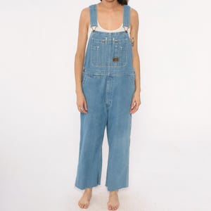 May include: Light blue denim overalls with adjustable straps, multiple pockets, and a relaxed fit. The overalls have a classic design with a button-fly closure and a small brown patch on the chest pocket. The denim is a medium wash, and the legs are cropped.