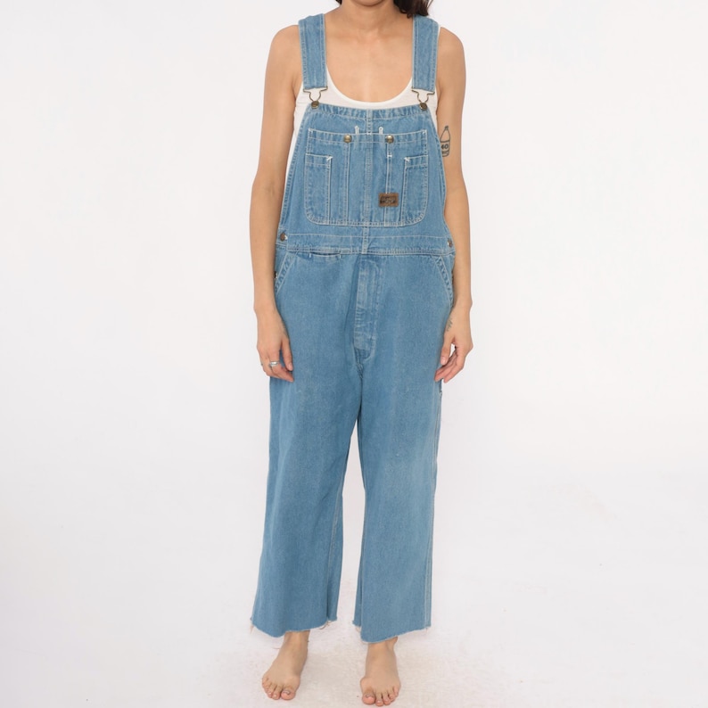 May include: Light blue denim overalls with adjustable straps, multiple pockets, and a relaxed fit. The overalls have a classic design with a button-fly closure and a small brown patch on the chest pocket. The denim is a medium wash, and the legs are cropped.