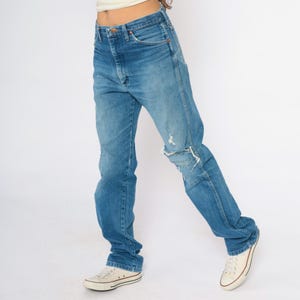 May include: Blue denim jeans with a distressed knee. The jeans have a classic five-pocket design and a high waist. The person is wearing white canvas trainers. The jeans are a medium wash and appear vintage.