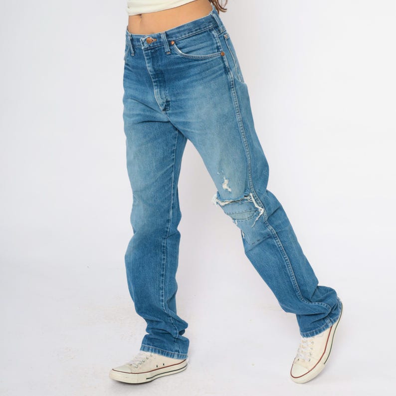 May include: Blue denim jeans with a distressed knee. The jeans have a classic five-pocket design and a high waist. The person is wearing white canvas trainers. The jeans are a medium wash and appear vintage.