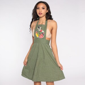 May include: Olive green apron dress with a patchwork apple design on the bib. The dress features a flared skirt and adjustable straps. The apple design includes red, yellow, and green fabric pieces.