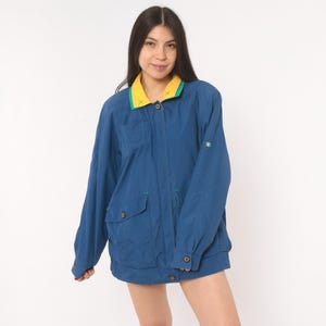 May include: A long-sleeve blue jacket featuring a contrasting yellow and green collar, a button-up front closure, two front flap pockets with buttons, and buttoned cuffs. The jacket has a relaxed fit and a solid blue color throughout the body.