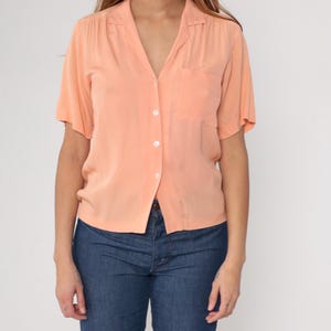 Puede incluir: Camisa melocot&oacute;n de manga corta con bolsillo en el pecho izquierdo. La camisa tiene cuello en V y se combina con vaqueros azules. La camisa est&aacute; abotonada y parece estar hecha de una tela ligera.