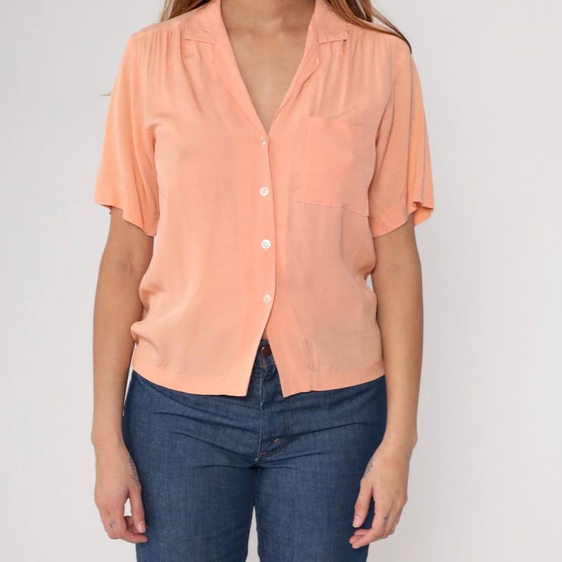 Puede incluir: Camisa melocot&oacute;n de manga corta con bolsillo en el pecho izquierdo. La camisa tiene cuello en V y se combina con vaqueros azules. La camisa est&aacute; abotonada y parece estar hecha de una tela ligera.