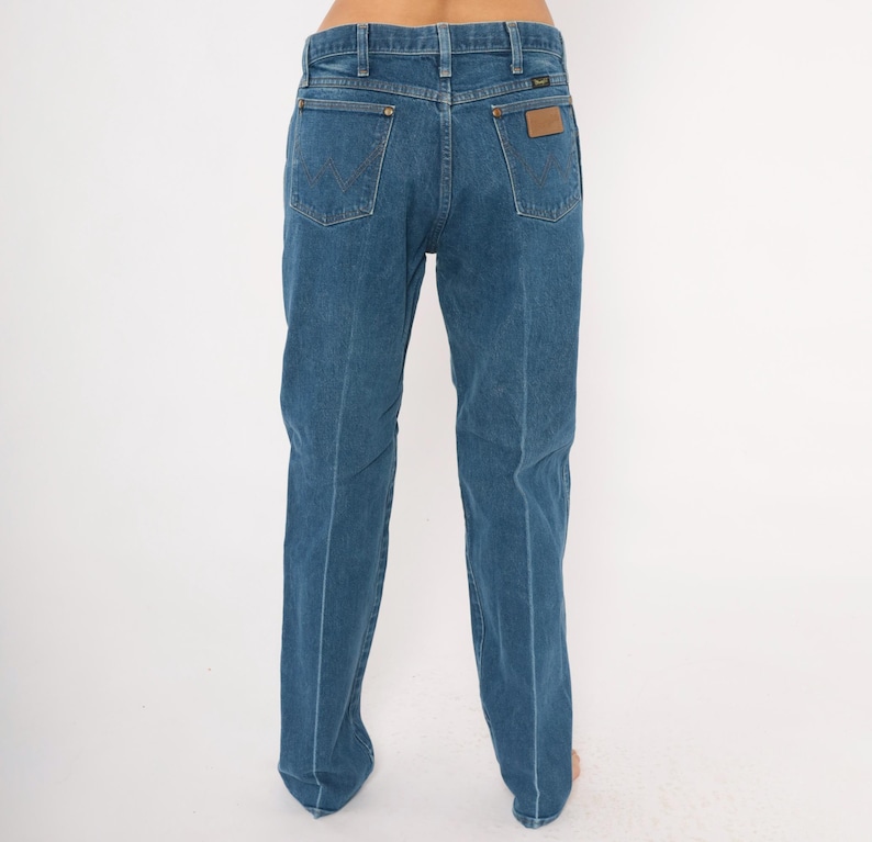Puede incluir: Vaqueros azules de mezclilla con un dise&ntilde;o cl&aacute;sico de cinco bolsillos. Los vaqueros tienen un corte de pierna recta y un parche de cuero marr&oacute;n en el bolsillo trasero. La marca Wrangler es visible.