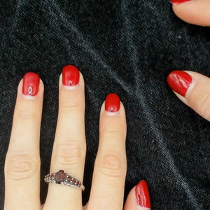 May include: Close-up of hands with red nail polish and a silver ring with a dark red stone. The nails are painted a vibrant red colour. The background is a dark, textured fabric.
