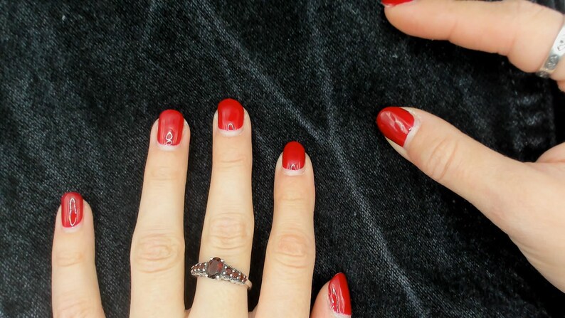 May include: Close-up of hands with red nail polish and a silver ring with a dark red stone. The nails are painted a vibrant red colour. The background is a dark, textured fabric.