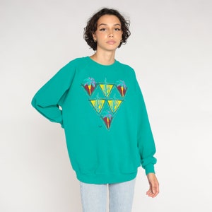 May include: Teal-colored sweatshirt with a triangular design featuring palm trees and cacti in shades of purple, yellow, and green. The long-sleeved top has a crew neck and a relaxed fit, suitable for casual wear.