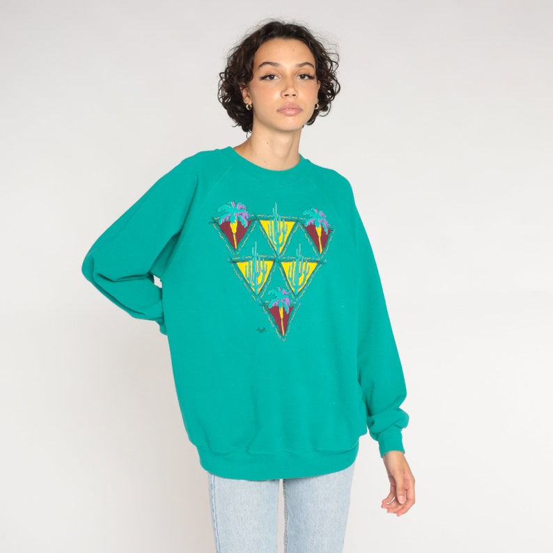 May include: Teal-colored sweatshirt with a triangular design featuring palm trees and cacti in shades of purple, yellow, and green. The long-sleeved top has a crew neck and a relaxed fit, suitable for casual wear.