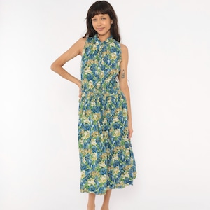 Op de afbeelding: Een mouwloze midi-jurk met een levendig bloemenpatroon in blauwe, groene en crèmekleurige tinten. Het kledingstuk heeft een kraag, een knoopsluiting aan de voorzijde en een geplooide taille die uitloopt in een wijde, geplooide rok.
