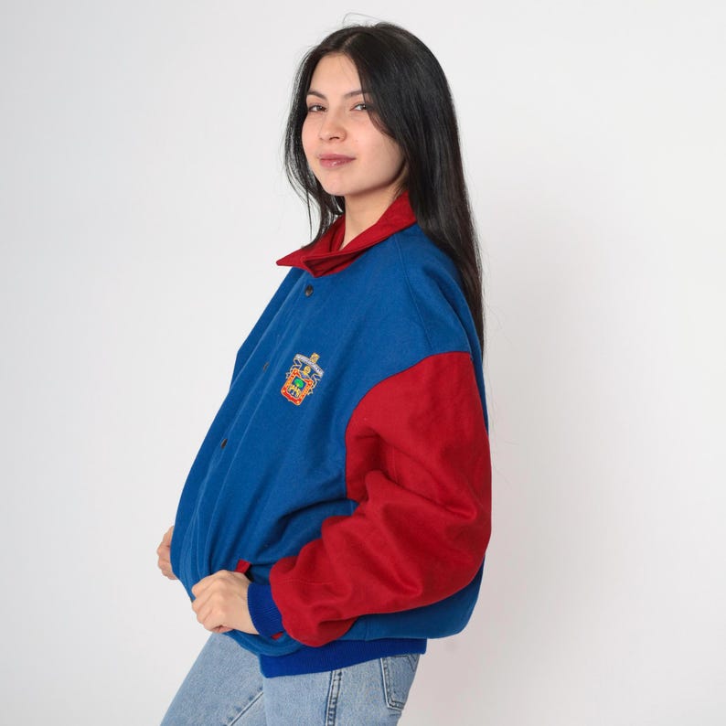 May include: A blue and red varsity jacket with a colorful embroidered logo on the left chest. The jacket has a snap closure and a ribbed collar, cuffs, and hem.