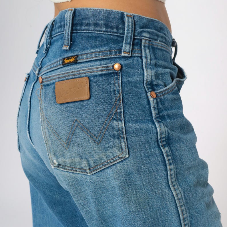 May include: Close-up of blue denim jeans with a classic design. The jeans feature a brown leather patch with the brand name and copper rivets. The back pocket has a distinctive stitching pattern. The jeans are a medium wash.