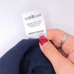May include: A white fabric garment care label attached to a dark blue textile. The label features black laundry symbols, text reading "Wash with similar colours, Reshape whilst damp, Do not iron motif," and the text "MADE IN THAILAND" at the bottom.