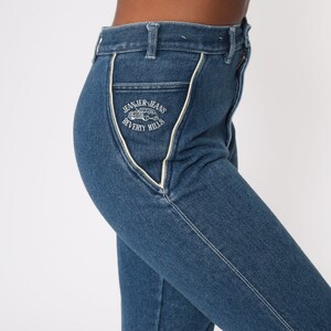 Puede incluir: Primer plano de unos vaqueros de mezclilla azules con cintura alta y pernera recta. Los vaqueros presentan costuras blancas y un ribete blanco a lo largo de los bolsillos. El bolsillo tiene un gr&aacute;fico blanco que dice "JEANIER-JEANS BEVERLY HILLS".