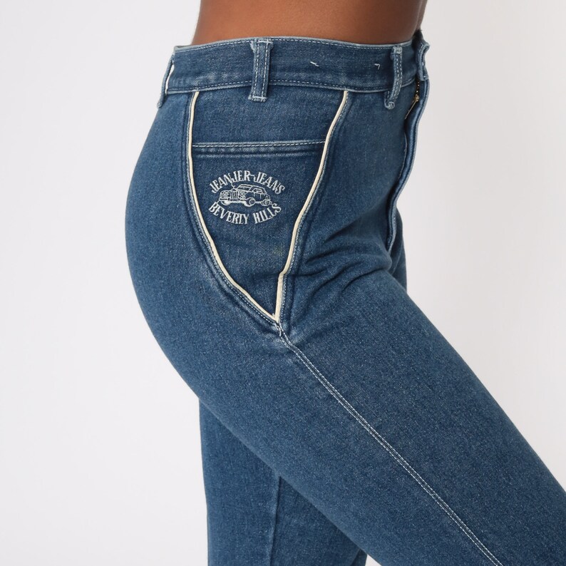 Puede incluir: Primer plano de unos vaqueros de mezclilla azules con cintura alta y pernera recta. Los vaqueros presentan costuras blancas y un ribete blanco a lo largo de los bolsillos. El bolsillo tiene un gr&aacute;fico blanco que dice "JEANIER-JEANS BEVERLY HILLS".