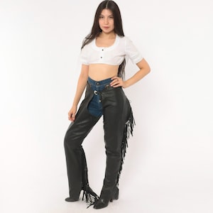 May include: A white short-sleeved crop top with a button-up front and lace trim on the sleeves, paired with dark blue denim jeans and black leather chaps featuring long fringe detailing along the outer legs and a silver buckle closure at the waist.