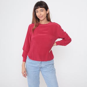 May include: A red long-sleeve blouse with a round neckline. The blouse has a relaxed fit and is paired with light blue jeans. The sleeves are slightly gathered at the cuffs. The model is standing in front of a white background.