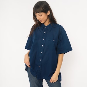 May include: A dark blue short-sleeved button-down shirt with two chest pockets. The shirt has a classic collar and is worn with blue jeans. The buttons are white, and the shirt appears to be made of a soft fabric.