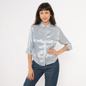May include: A light blue satin shirt with a collared neckline, two chest pockets, and three-quarter sleeves. The shirt is button-down and has a shiny, smooth texture. The model is wearing blue jeans.