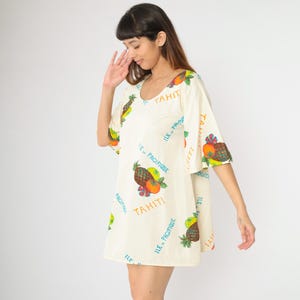 Pode incluir: Um vestido branco de manga curta com estampa de frutas tropicais e o texto "Tahiti, Ile-Pacifique" repetido em toda a superfície.