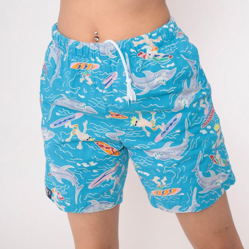 K&ouml;nnte beinhalten: Blaue Badehose mit einem bunten Druck von Haien, Surfern und Wellen.
