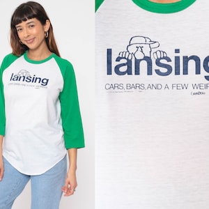 May include: A white and green baseball tee shirt with a blue and white graphic of a cartoon character peeking over the text "lansing" and the words "Cars, Bars, and a Few Weirdos".
