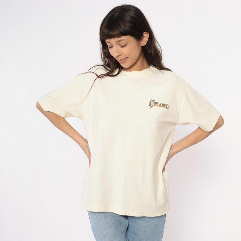 May include: Cream-colored t-shirt with short sleeves and a mock neck. The shirt features the text "GIRL SCOUTS" in a gold and green design on the upper right chest. The model is wearing blue jeans.