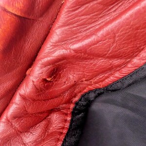 May include: Close-up of red leather material with visible wear and tear. The leather has a textured surface and shows signs of damage, including a small tear. A black fabric is visible along the edge, with a black stitched seam.