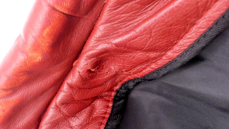 May include: Close-up of red leather material with visible wear and tear. The leather has a textured surface and shows signs of damage, including a small tear. A black fabric is visible along the edge, with a black stitched seam.