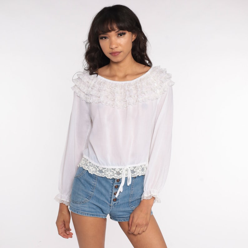 May include: White long-sleeved blouse with a ruffled neckline and lace trim. The blouse has a cinched waist and is made of a lightweight fabric.