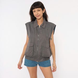 May include: A charcoal gray sleeveless utility vest featuring a full-length front zipper, a classic collar, and two large flap pockets at the waist. The garment includes a smaller vertical zippered pocket on the upper left chest and a ribbed hem.