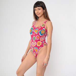 May include: A one-piece swimsuit in a vibrant pink hue, adorned with a colorful paisley and floral pattern. The swimsuit features a scoop neckline and high-cut leg openings, offering a retro aesthetic. The design incorporates various shapes and bright colors.