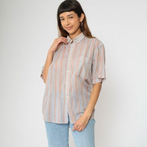 May include: A button-down shirt with vertical stripes in shades of gray and peach. The short-sleeved shirt has a chest pocket and is paired with light blue jeans. The shirt is made of a lightweight fabric.
